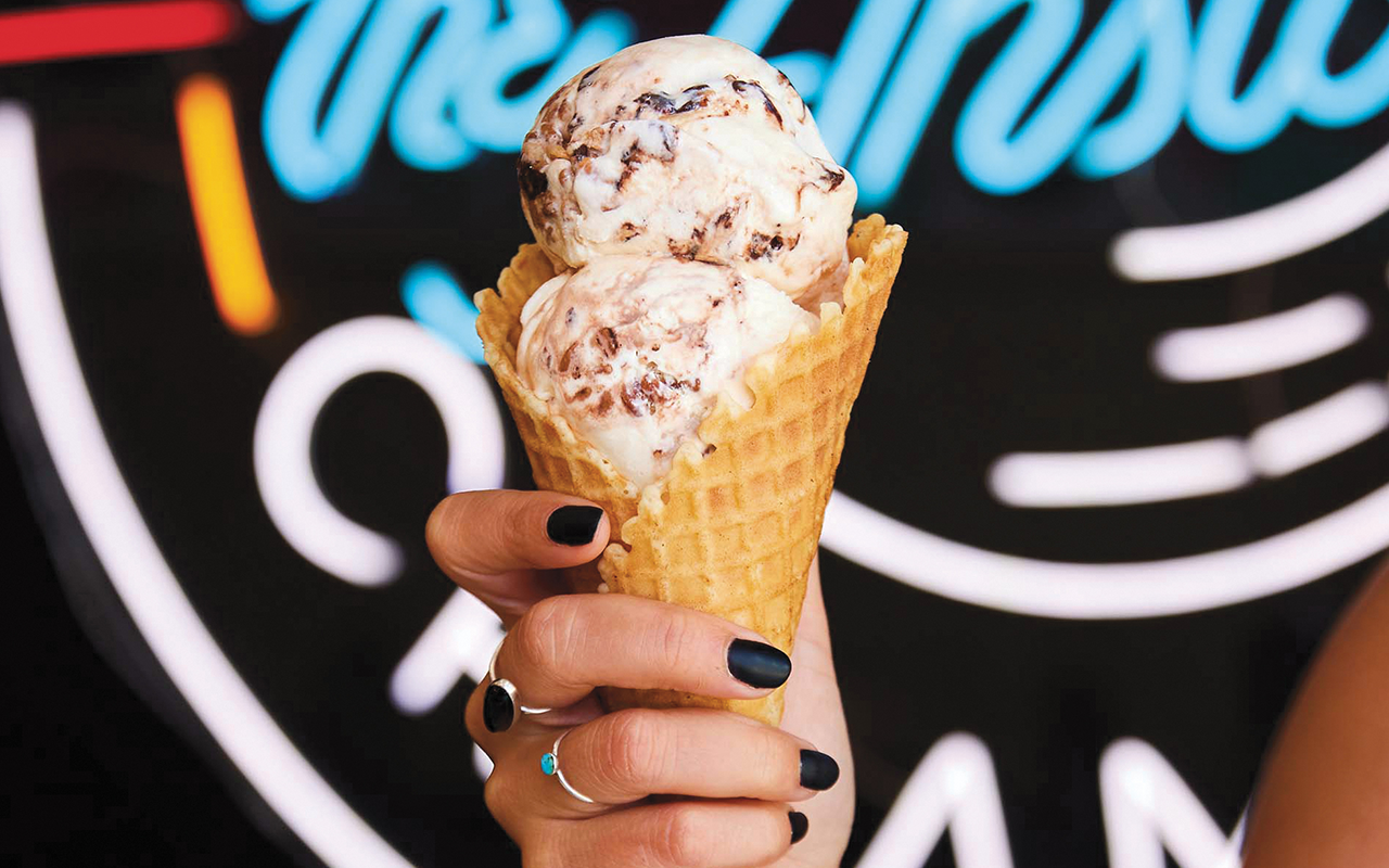 Ice cream cone held by a hand with dark nail polish in front of a neon sign