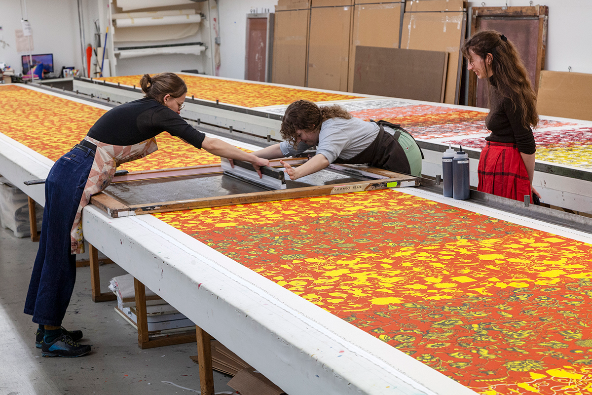 Artists working on a large silk screening tapestry in shades of orange and yellow