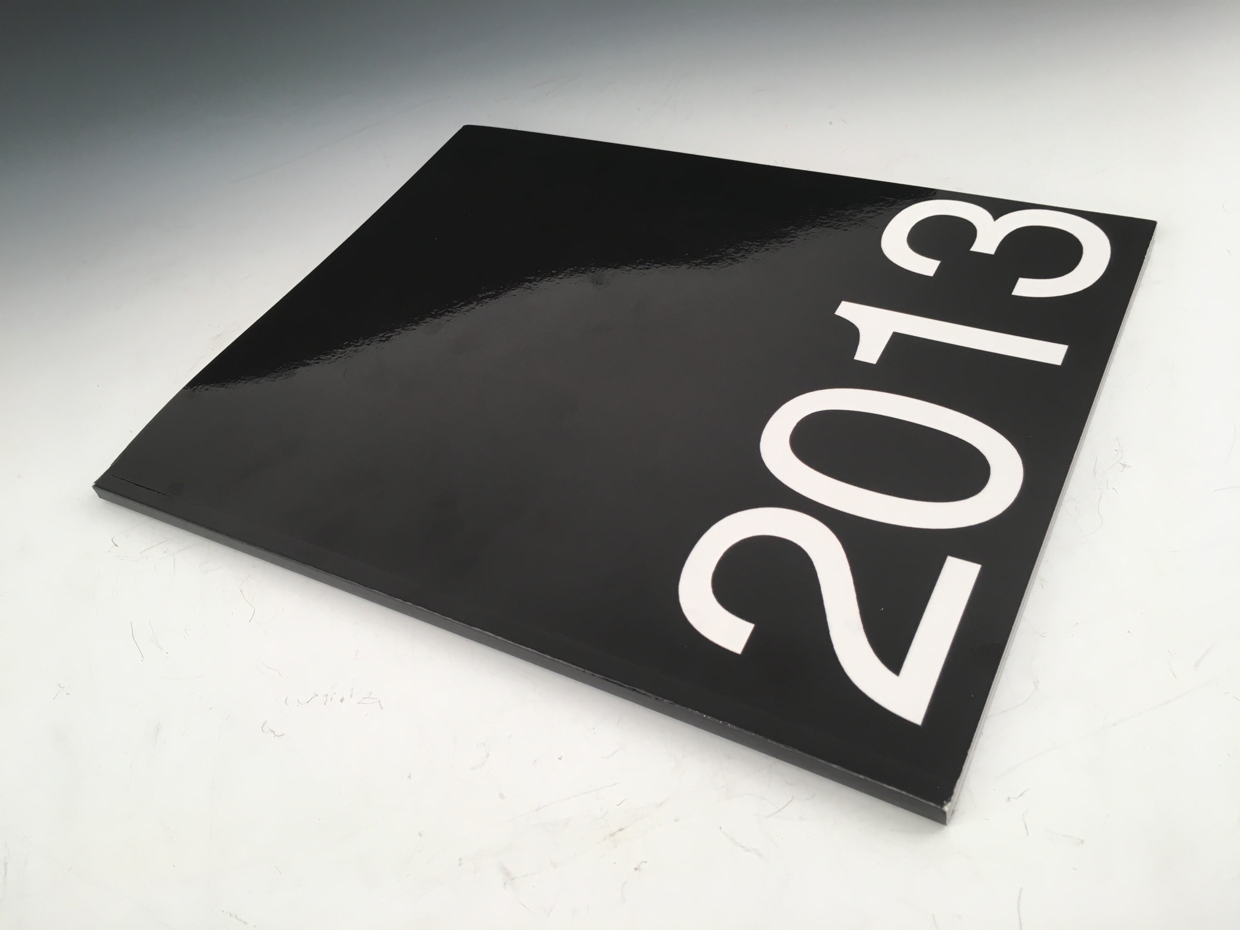 A photograph of 2013, 2023, a black glossy paperback art book, lying on a flat marble surface