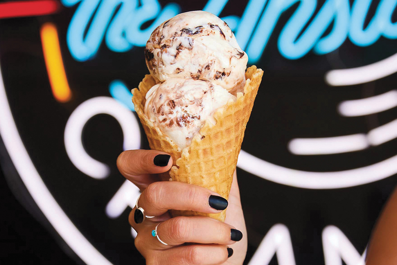 Ice cream cone held by a hand with dark nail polish in front of a neon sign